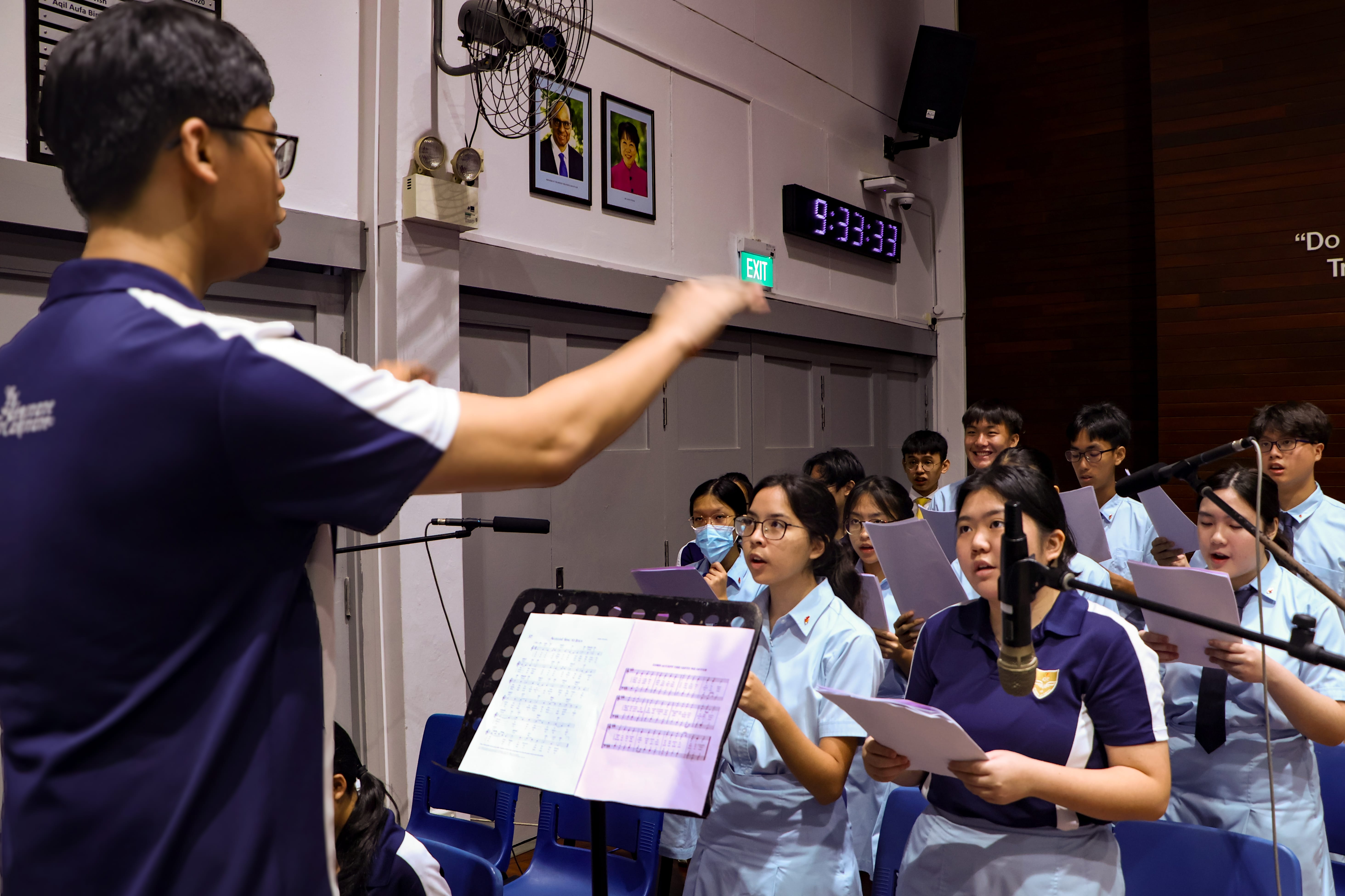 Conductor leads a student choir holding sheets in a hall, with microphones and a digital clock on the wall.